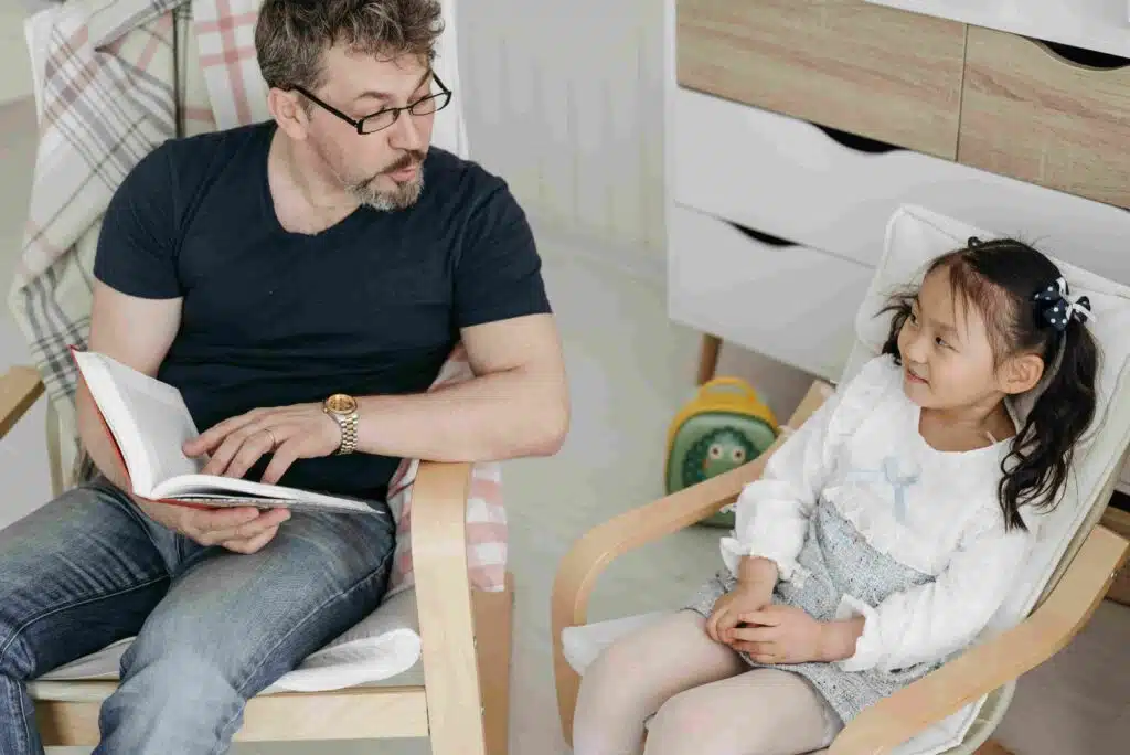 A man sits in a chair with a little girl, who is reading a book, in a cozy short-term care setting.